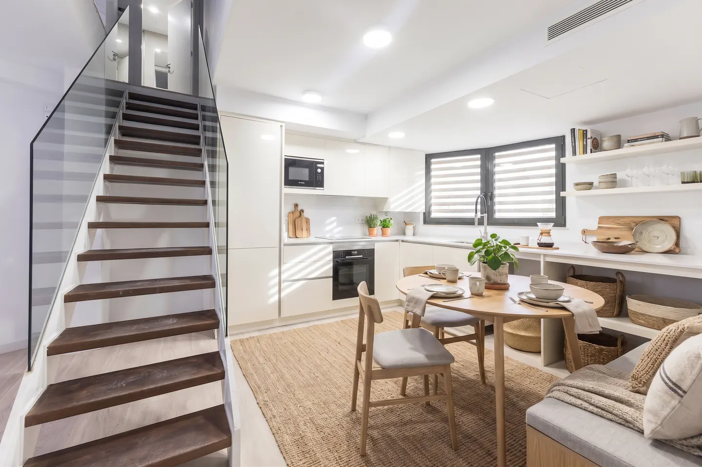 A bright, modern kitchen with white cabinets, a round wooden table with chairs, and a staircase with glass railing.