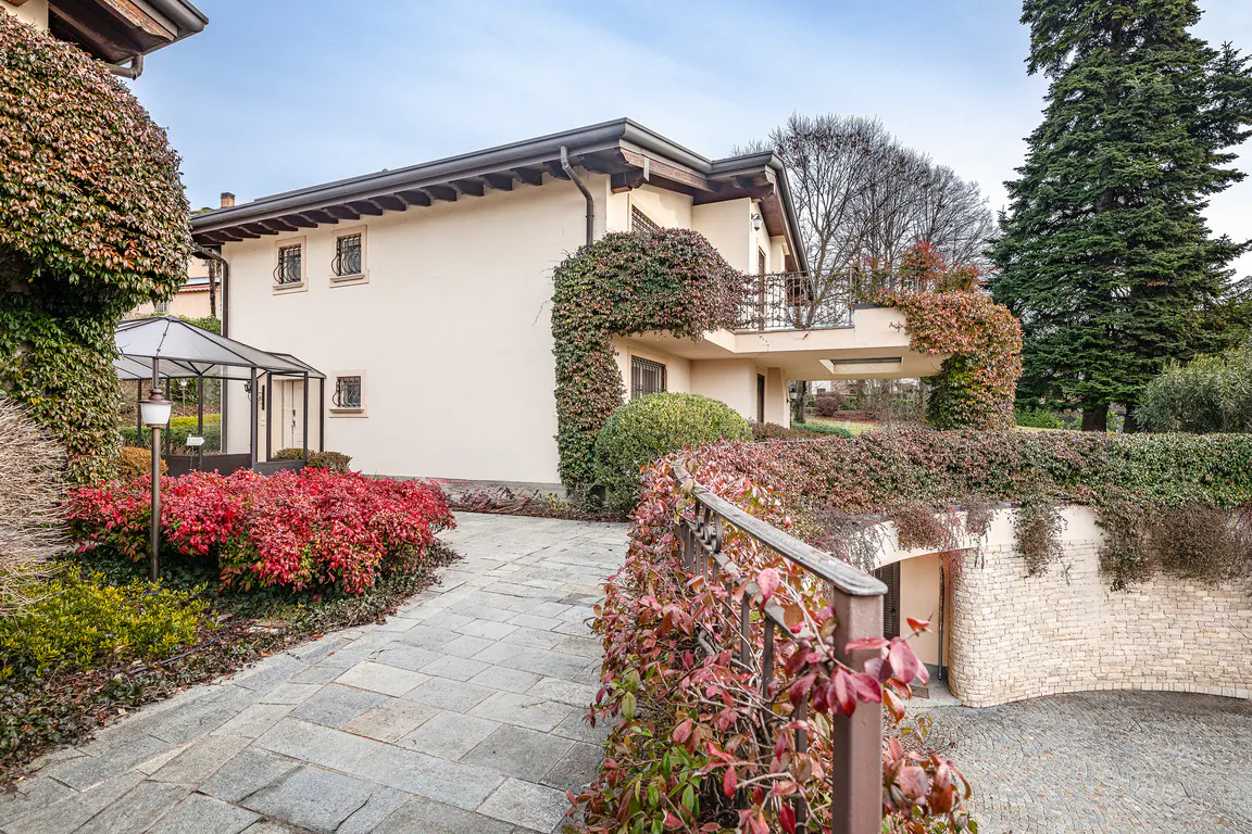 Two-story beige house with a stone walkway, red bushes, and green ivy climbing the walls. A stone wall and garage entrance are on the right.