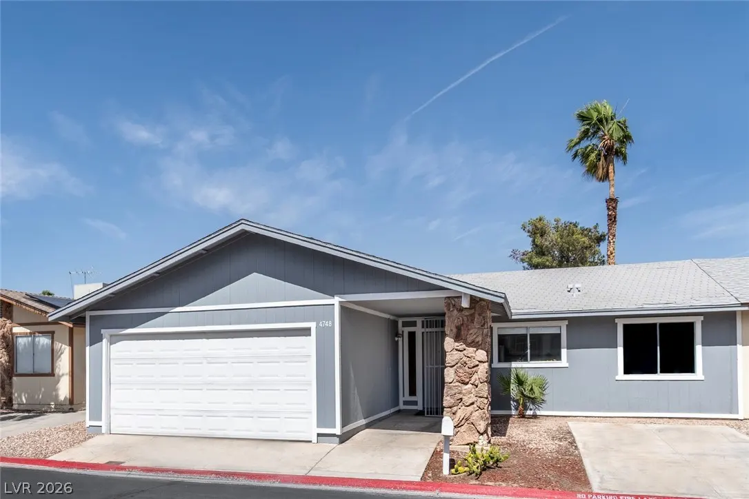 A gray single-story house with a white garage door under a blue sky. A palm tree is visible in the background.