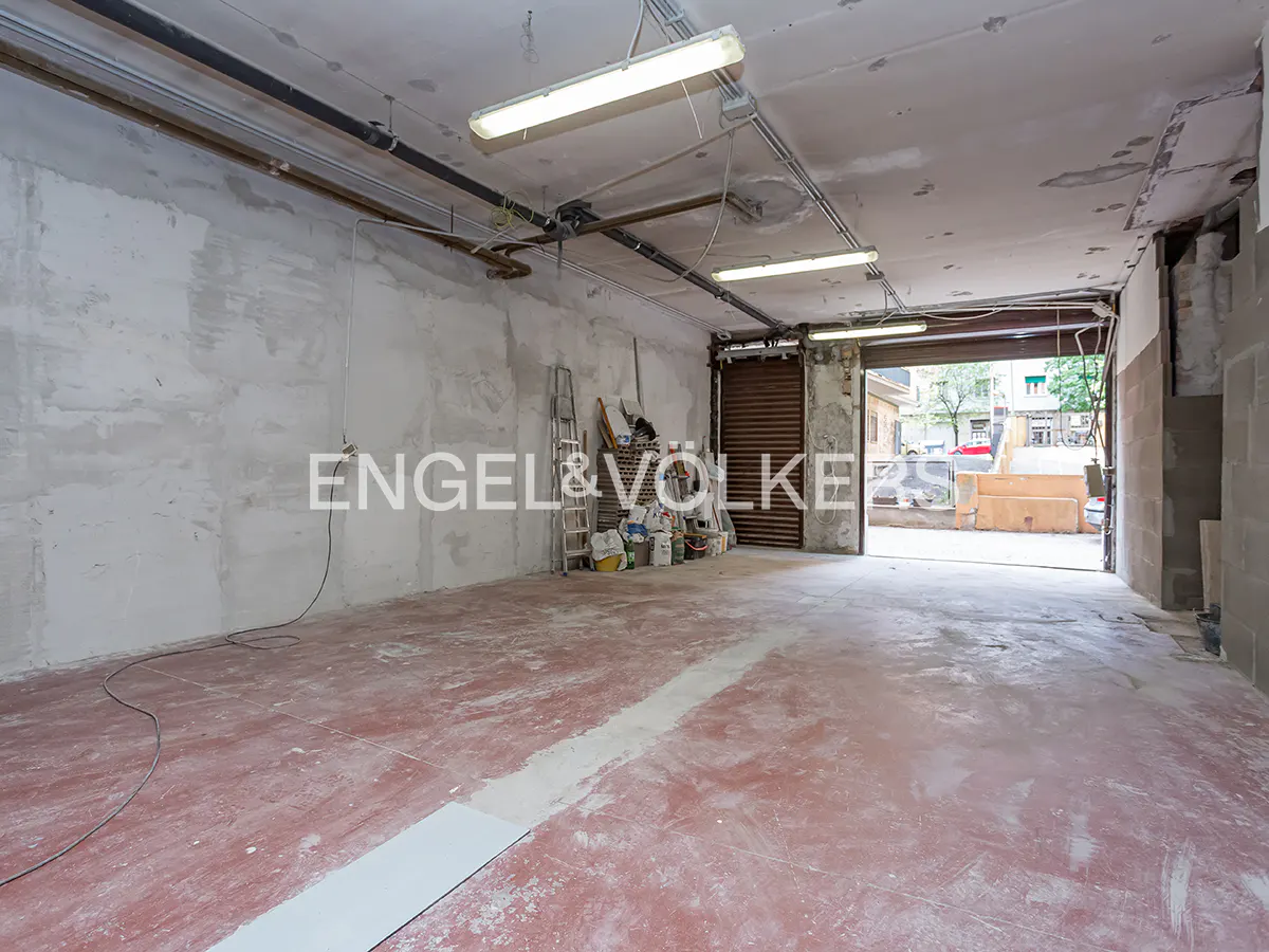 Unfinished commercial space with a red floor, gray walls, and an open garage door leading to a street view. Fluorescent lights illuminate the interior.