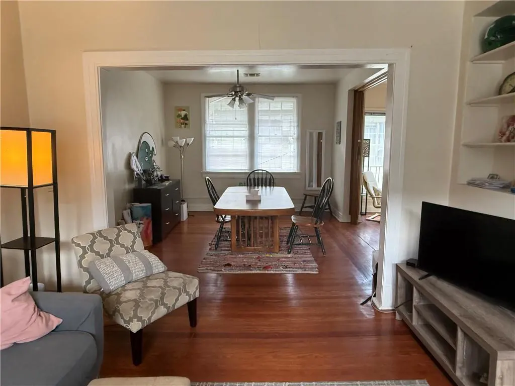 A living room with hardwood floors, a patterned chair, and a view into the dining room with a table and chairs. A TV is on the right.