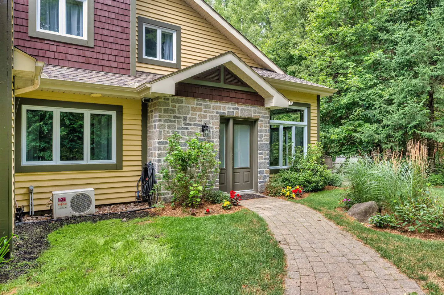 Exterior of a two-story home with yellow siding, brown shingles, and a stone entryway. A brick path leads to the front door. Green lawn and trees surround the house.