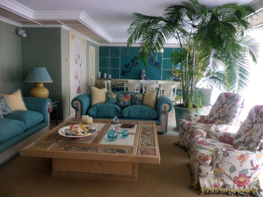 Living room with teal sofas, floral chairs, and a large palm tree. A wooden coffee table sits in the center.
