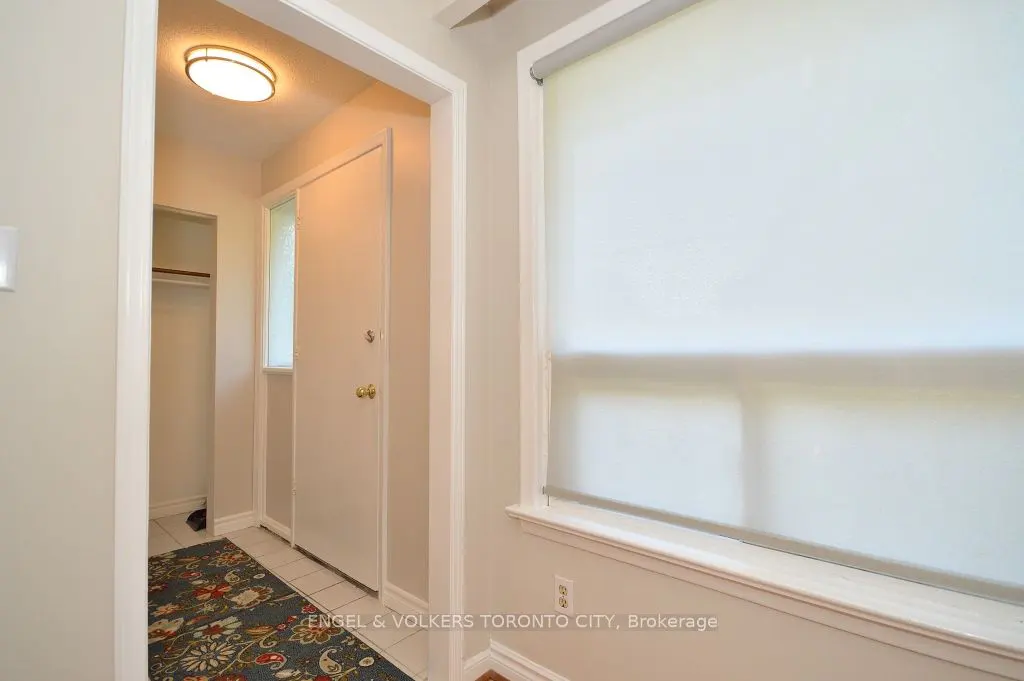 A bright foyer with a white door, closet, and a window with a white roller shade. A colorful rug is on the floor.