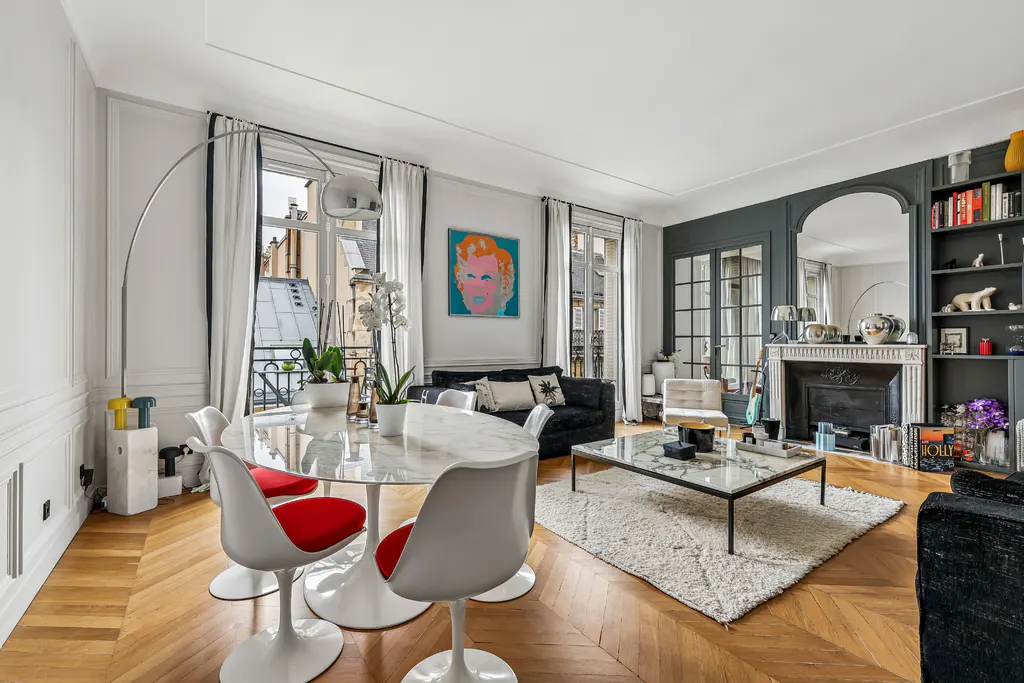 Bright living room with herringbone wood floors, white walls, and a marble table with red chairs. Windows overlook city buildings. Artwork and a fireplace add character.