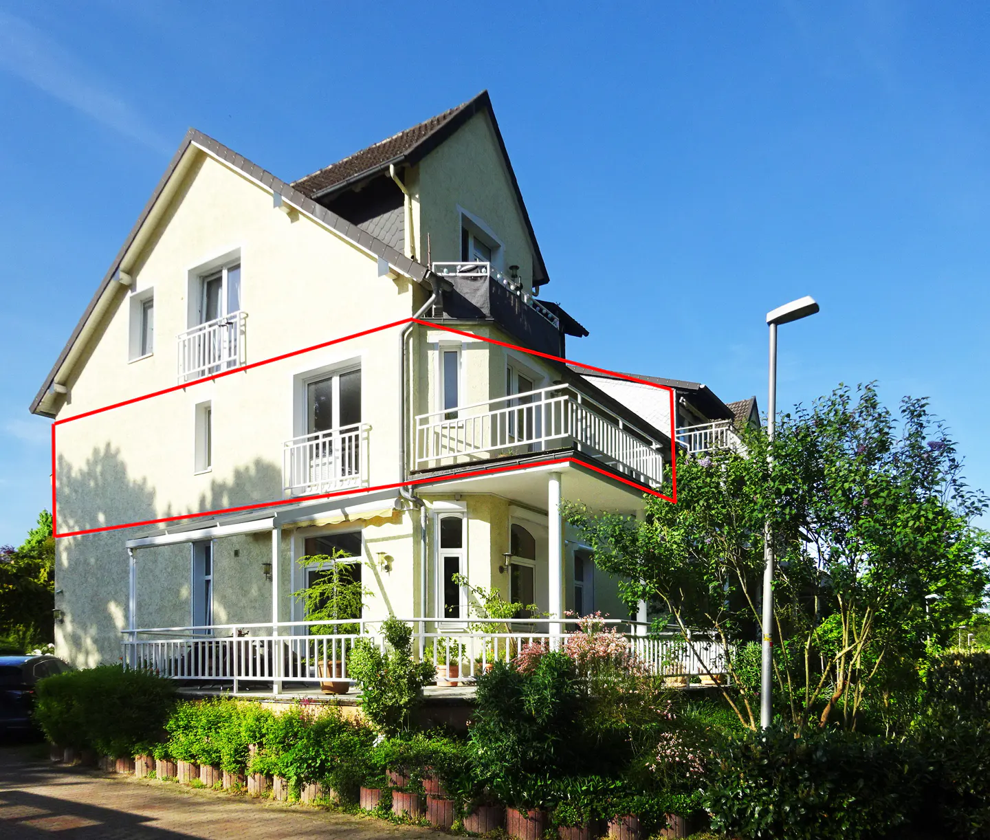 Two-story yellow house with white balconies and a brown roof under a clear blue sky. Lush green landscaping surrounds the property.