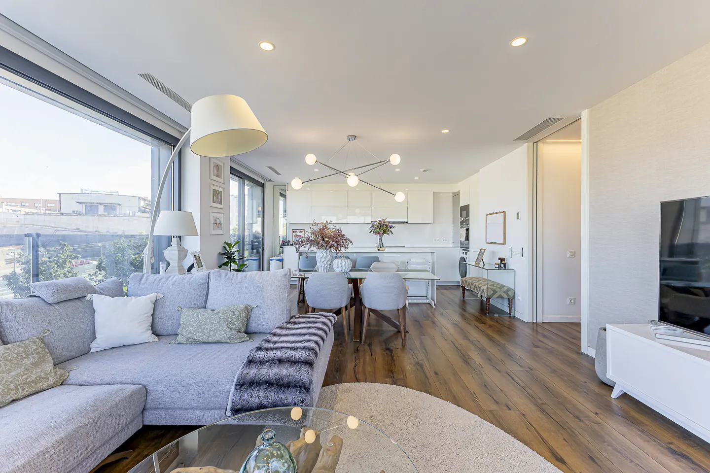 Bright, modern living room with a gray sectional sofa, wood floors, and a view from a large window. A dining area and kitchen are in the background.