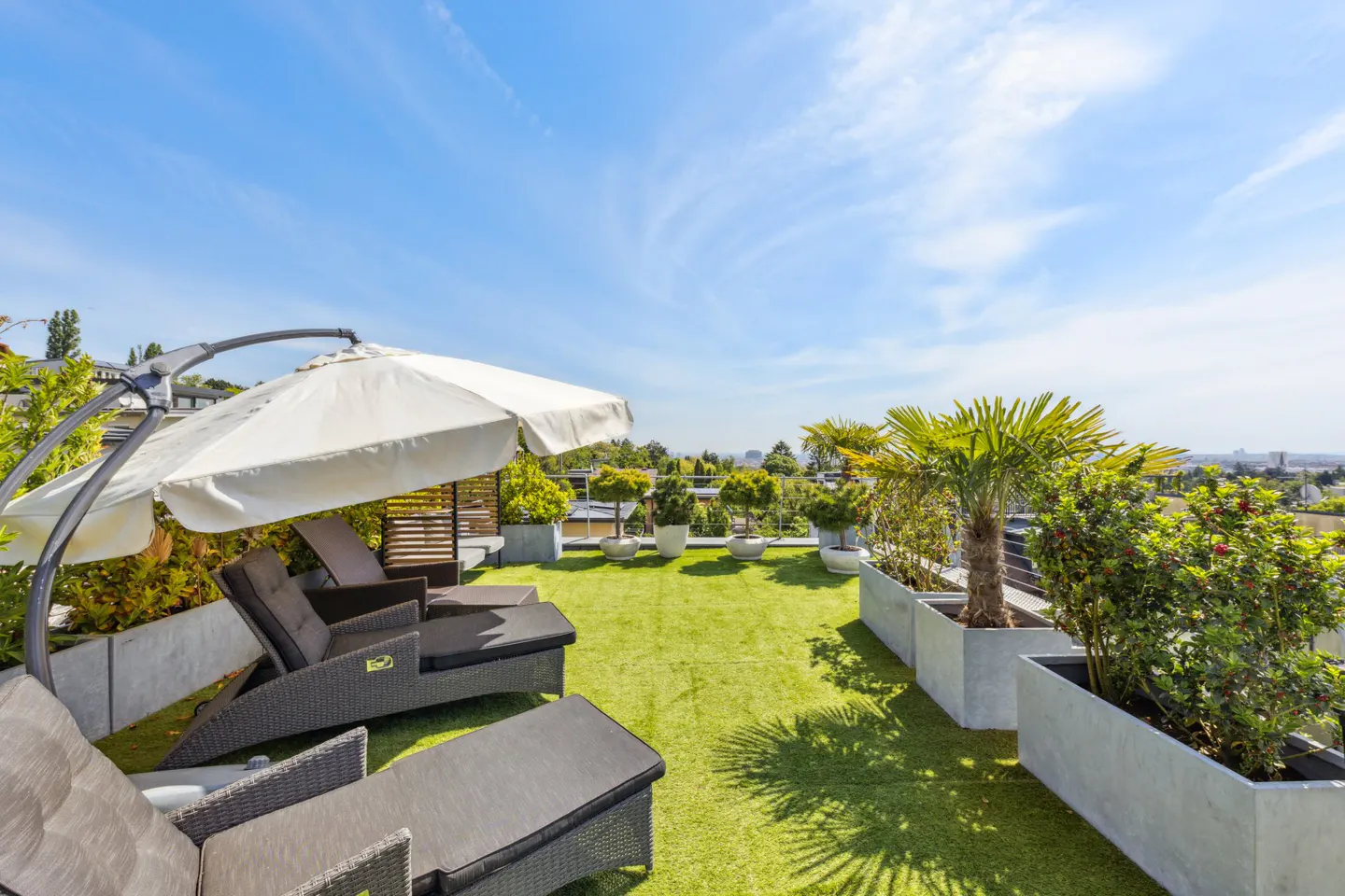 Sunny rooftop terrace with artificial grass, lounge chairs, a white umbrella, and potted plants under a clear blue sky.