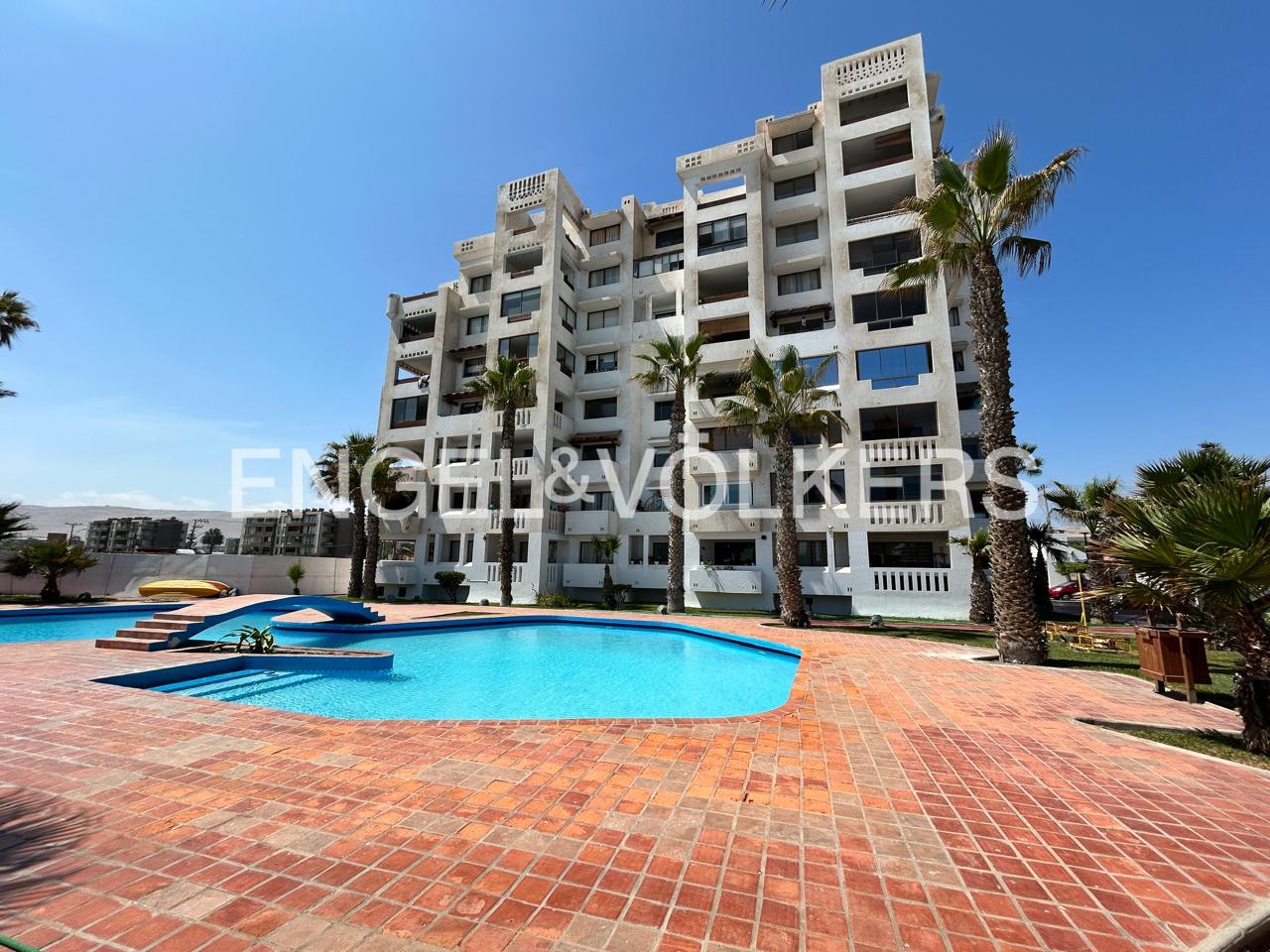 Multi-story white apartment building with balconies, a blue swimming pool, and palm trees on a sunny day.
