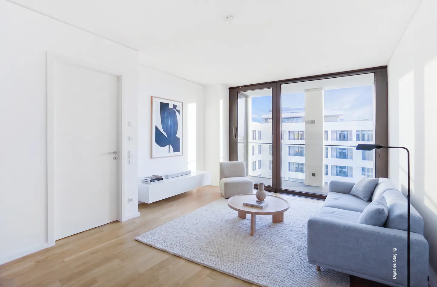 Bright living room with wood floors, white walls, and a large window. A blue sofa, chair, and round table sit on a light rug. A blue abstract art piece hangs on the wall.