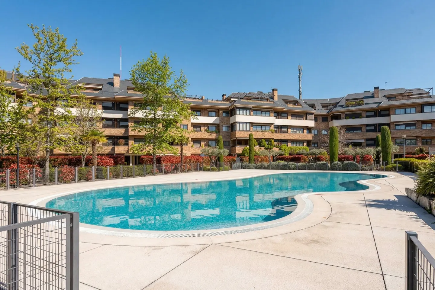 Outdoor pool with turquoise water in front of a brick apartment building under a clear blue sky.
