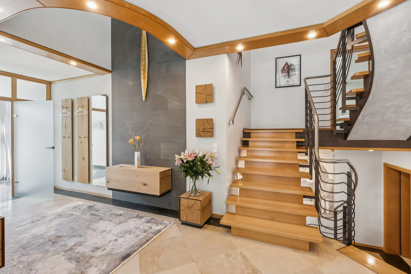 Bright foyer with wooden stairs, metal railings, and travertine floors. A gray rug and wooden furniture accent the space.