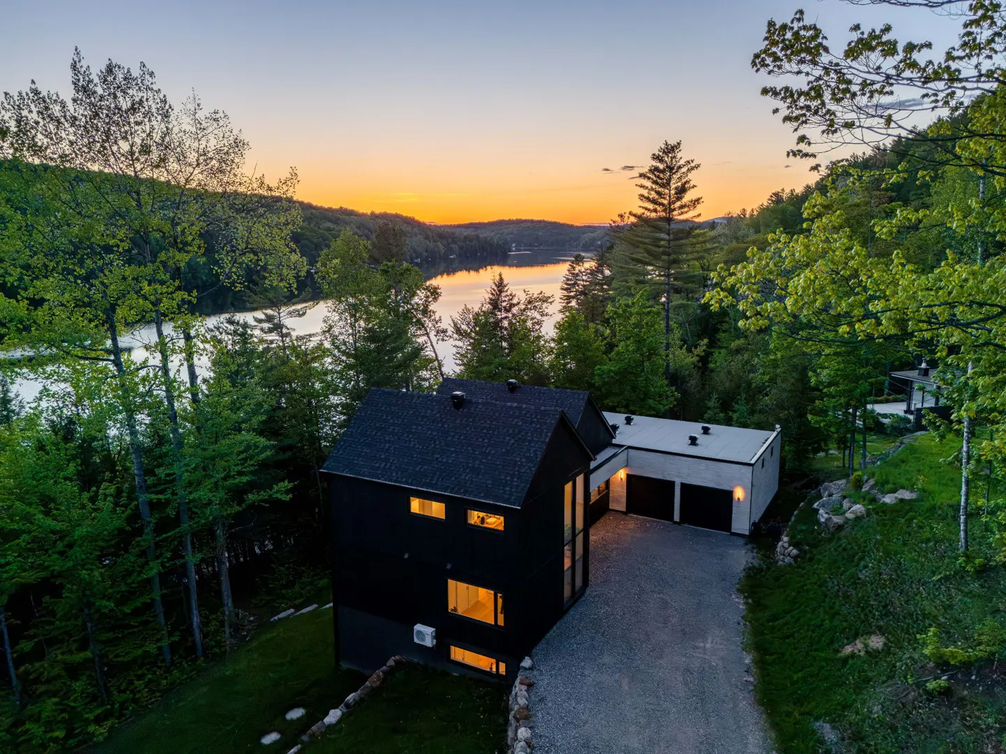 Modern black home with a gravel driveway overlooking a lake at sunset, surrounded by lush green trees.