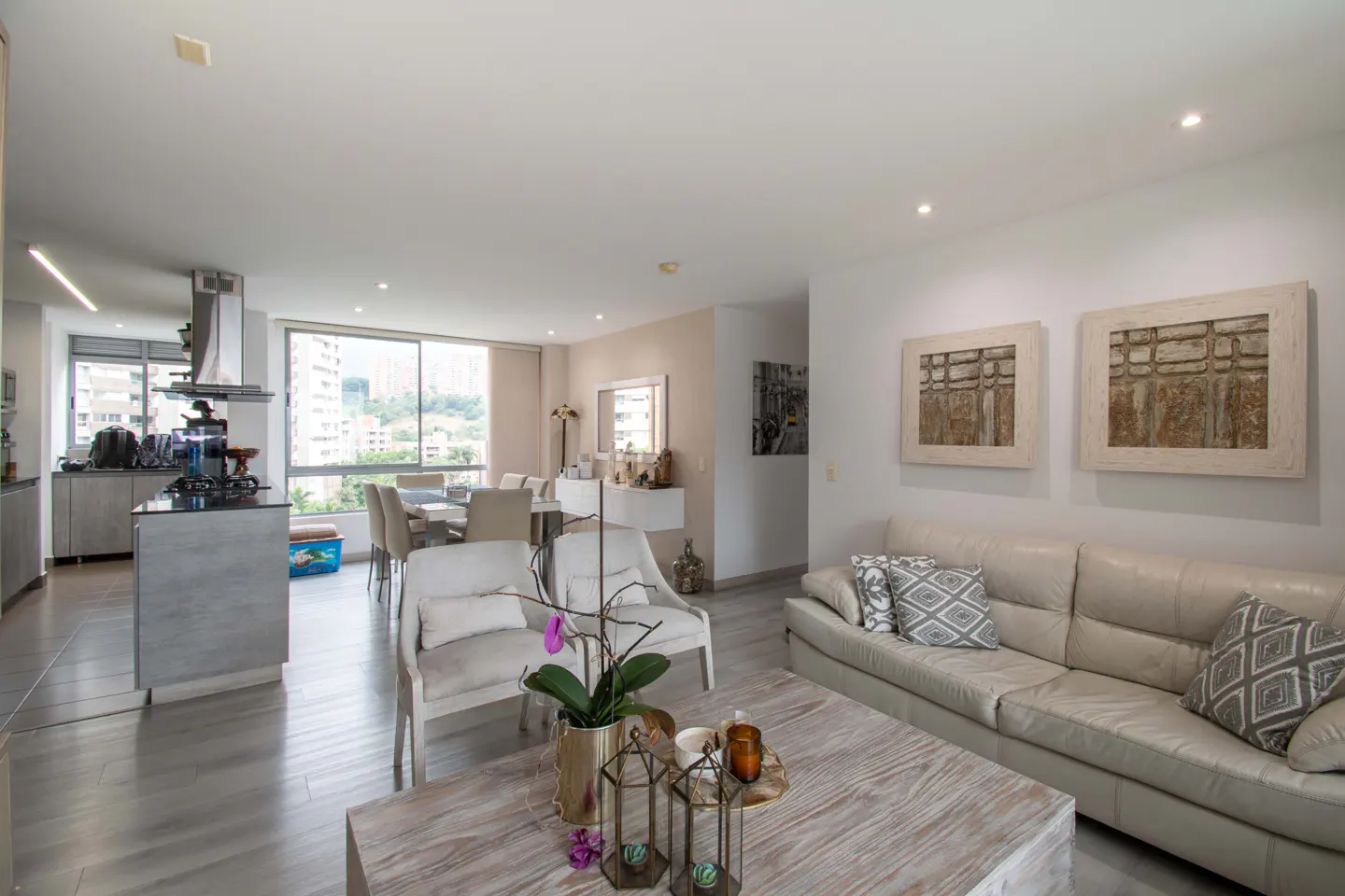 Bright, open-concept living space with a white sofa, wood coffee table, and dining area. Art adorns the walls. A kitchen island is visible.
