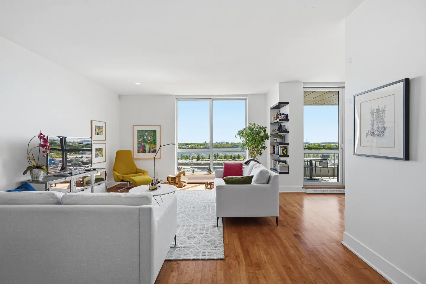 Bright living room with white sofas, wood floors, and a large window overlooking a river. A yellow chair and artwork add pops of color.