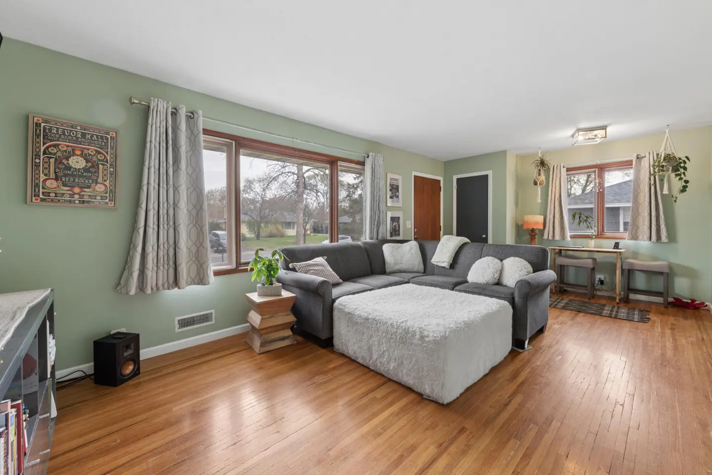 Living room with hardwood floors, green walls, and a gray sectional sofa with a white ottoman. Windows with curtains let in natural light.