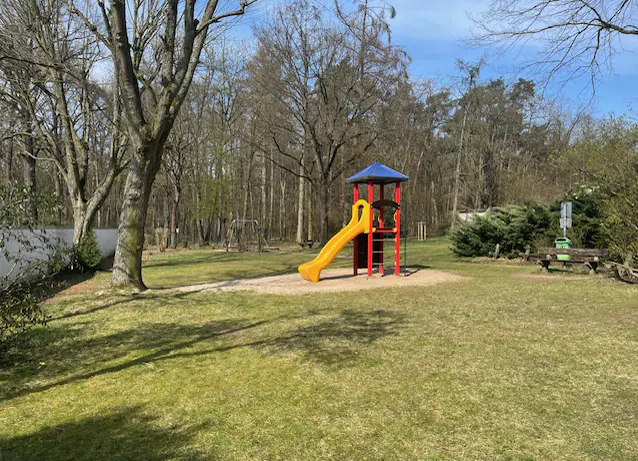 A playground with a yellow slide and blue roof sits on a grassy lawn surrounded by trees.