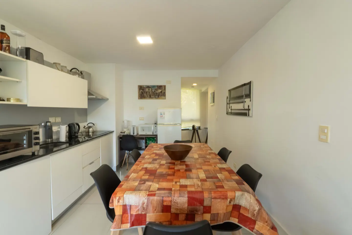 A bright kitchen with white walls, cabinets, and appliances. A dining table with a colorful tablecloth and black chairs is in the center.