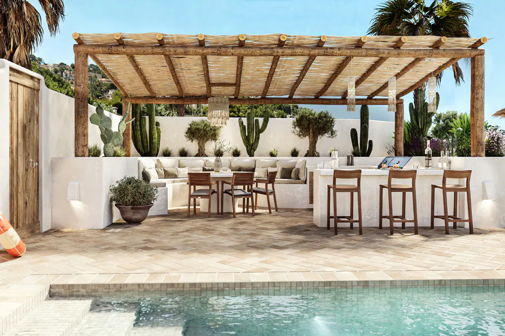 Outdoor patio with a pool, pergola, and bar area. Cacti and trees line the back wall. Table and chairs are set for dining.