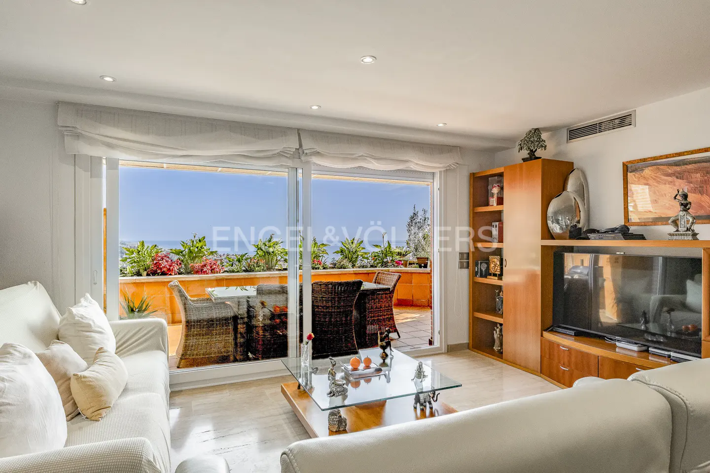 Bright living room with white sofas, a glass coffee table, and a balcony with ocean views. A wooden cabinet holds a TV and decorative items.