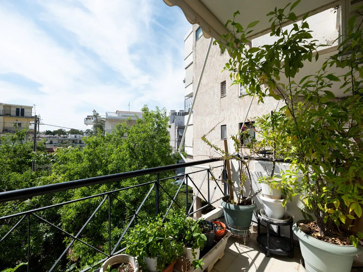 Balcony view with black iron railing, potted plants, and city buildings in the background under a blue sky with clouds.