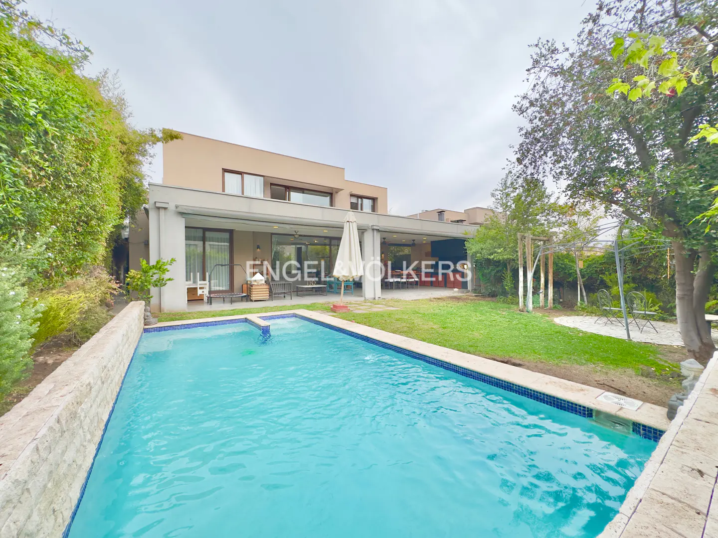 Modern two-story house with a backyard swimming pool, covered patio, and lush green landscaping under a cloudy sky.