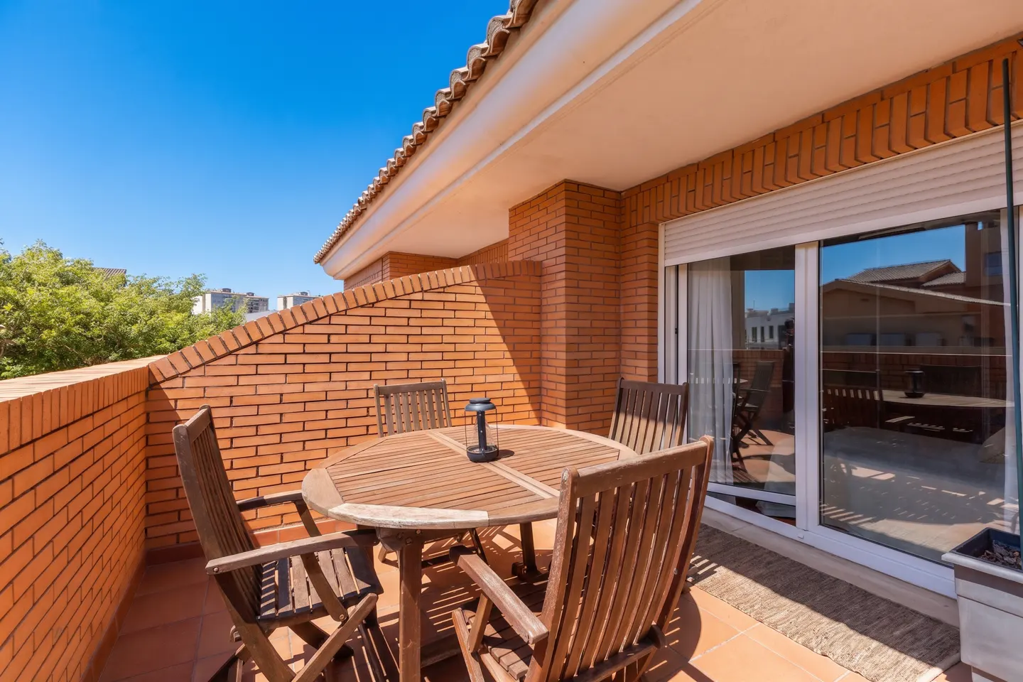 Outdoor patio with a round wooden table, four chairs, and a lantern. The patio has a brick wall and a sliding glass door.