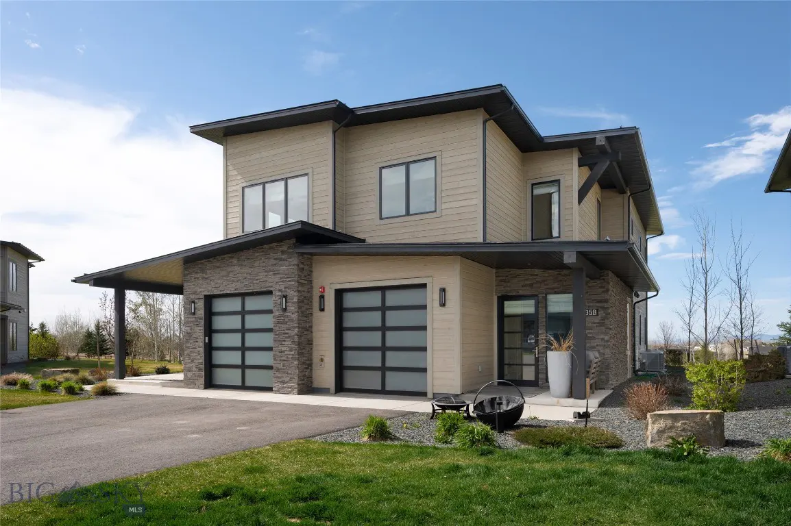 Two-story modern home with tan siding, stone accents, and black trim. Two-car garage with frosted glass doors. Green lawn and blue sky.