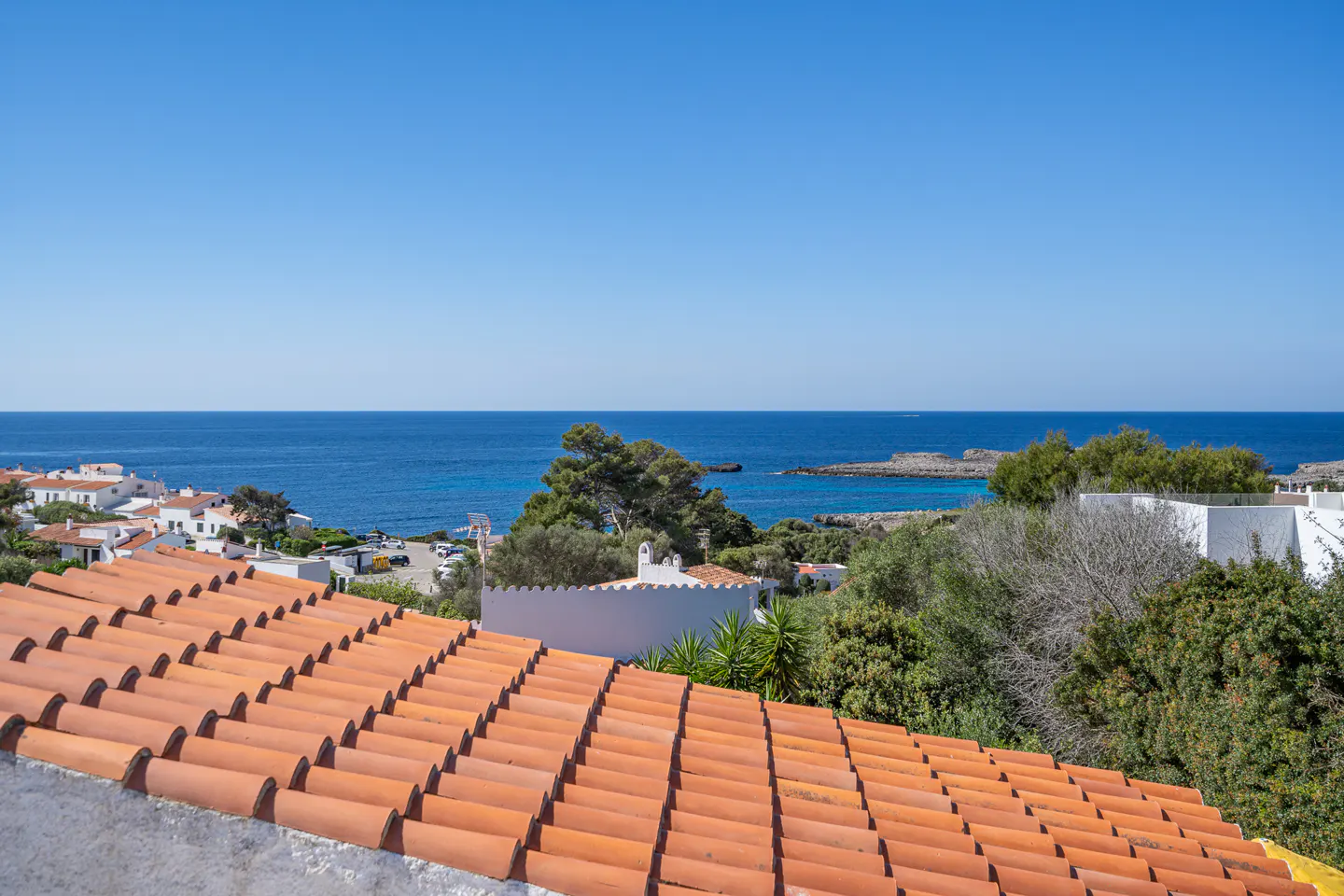 View of the ocean from a rooftop with orange tiles, with white buildings and green trees in the background under a clear blue sky.