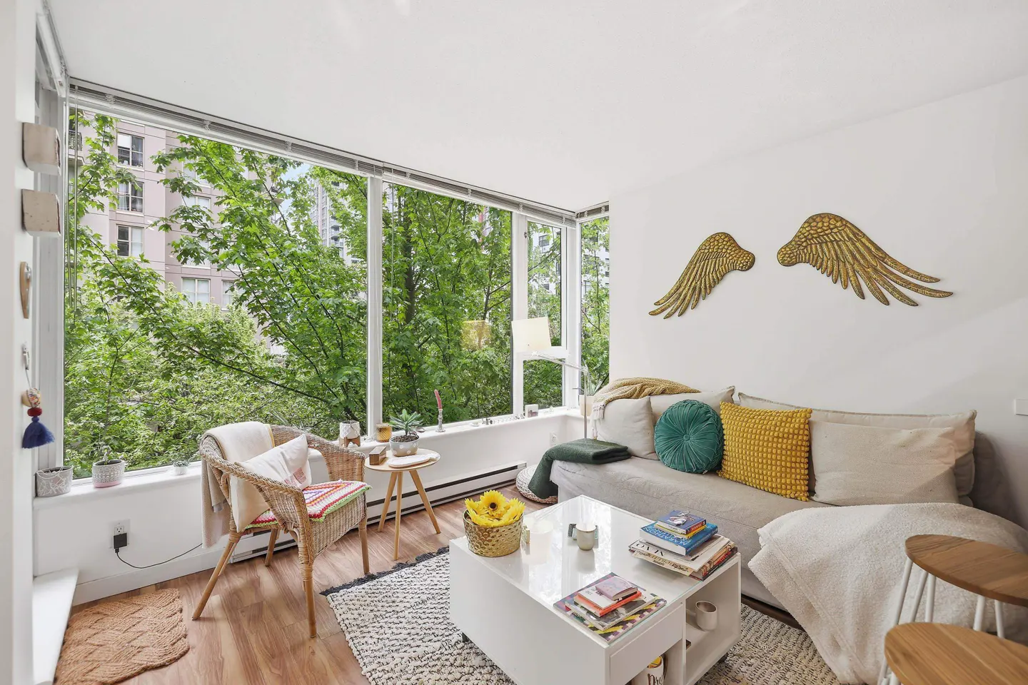 Bright living room with a large window view of trees. A sofa with pillows, a white coffee table, and gold wings on the wall.