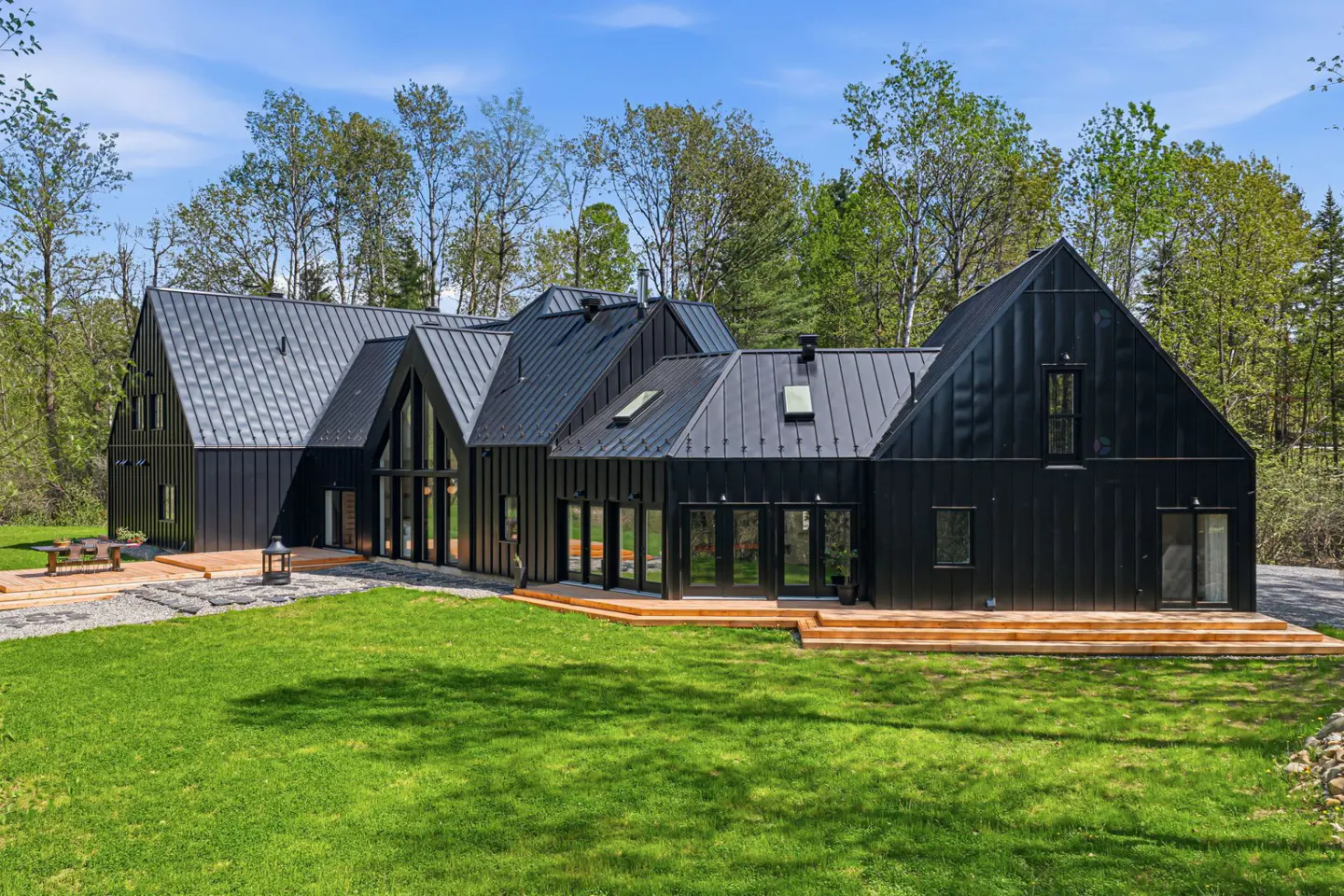 Modern black farmhouse with a metal roof, surrounded by green lawn and trees under a blue sky.