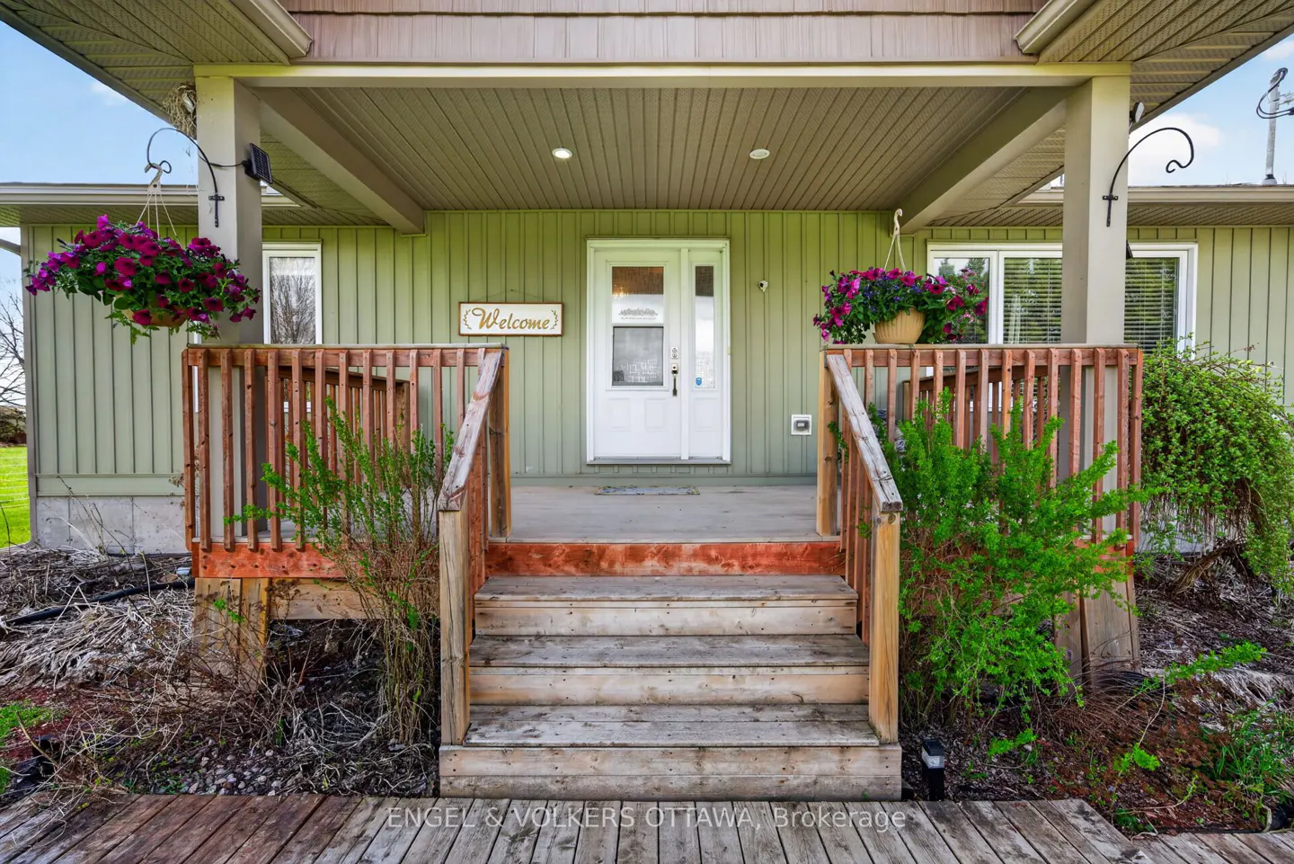 Front porch of a light green house with white door, wooden steps, and hanging flower baskets. A "Welcome" sign is above the door.