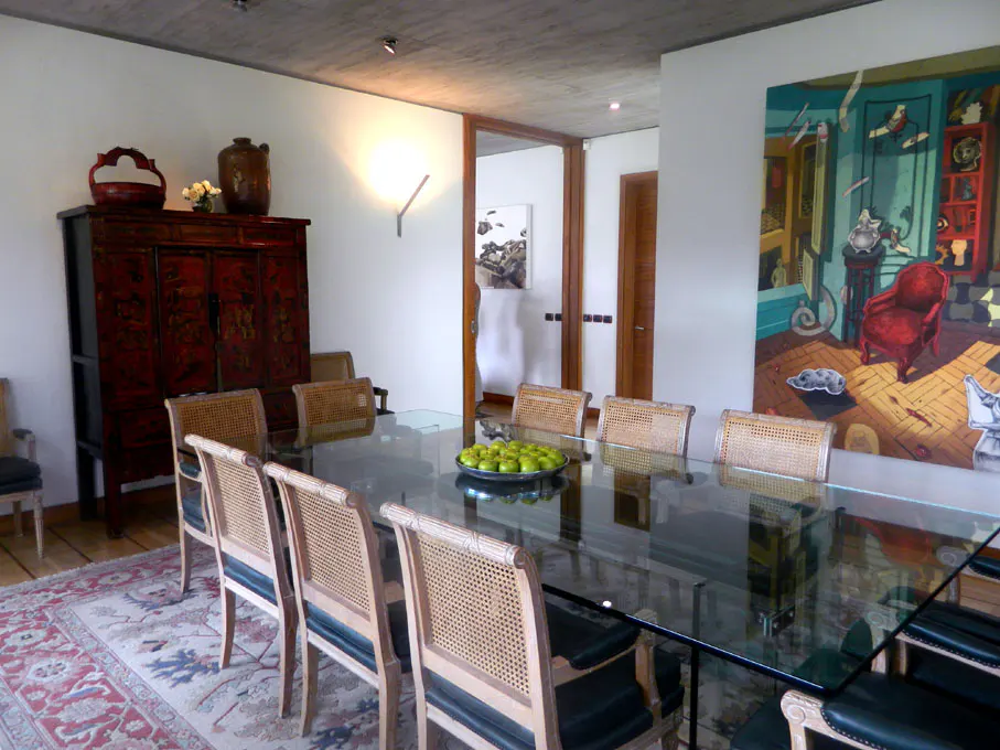 A dining room with a glass table, wicker chairs, and a red oriental cabinet. A colorful painting hangs on the wall.