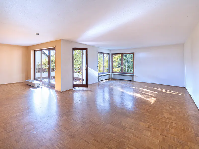 Bright, empty room with parquet floors, white walls, and large windows. Sunlight streams in, highlighting the open space and view of greenery outside.