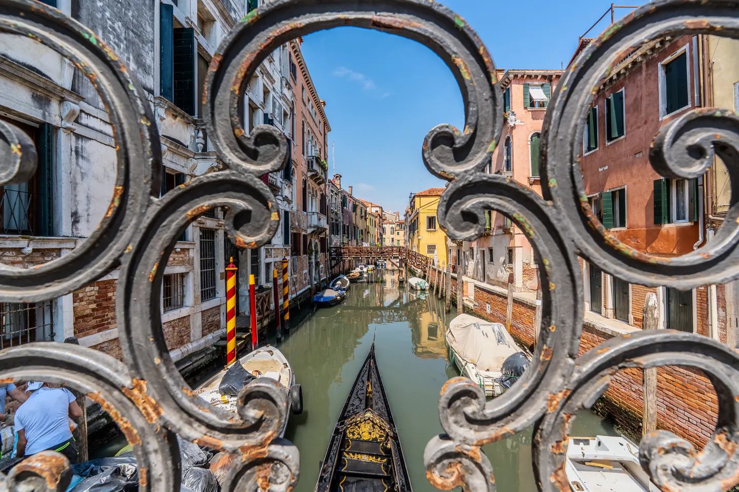 View of a Venice canal with a gondola and boats, framed by an ornate, weathered black iron bridge railing.