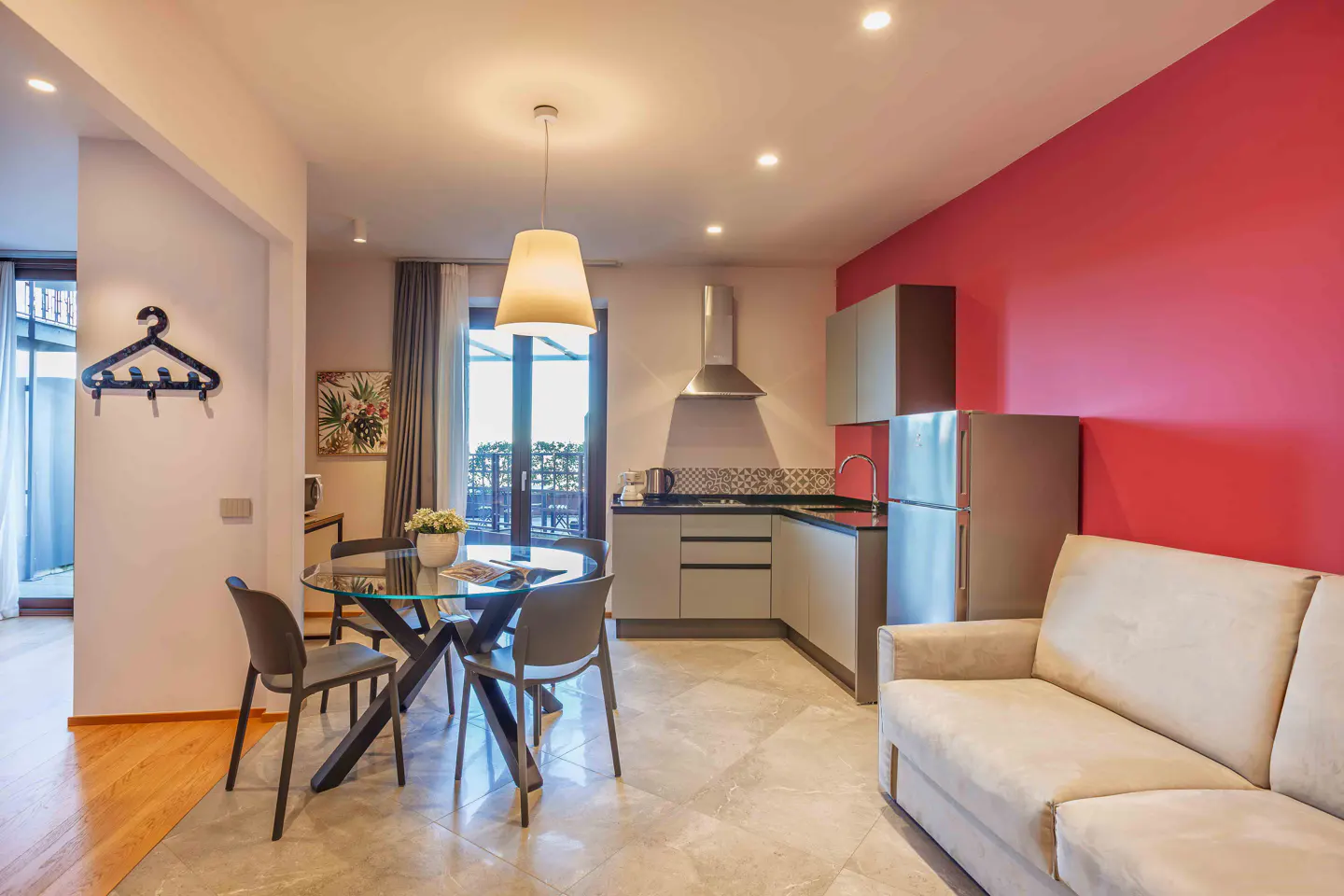 Bright apartment interior with a glass table, grey chairs, and a kitchen with stainless steel appliances against a red wall.