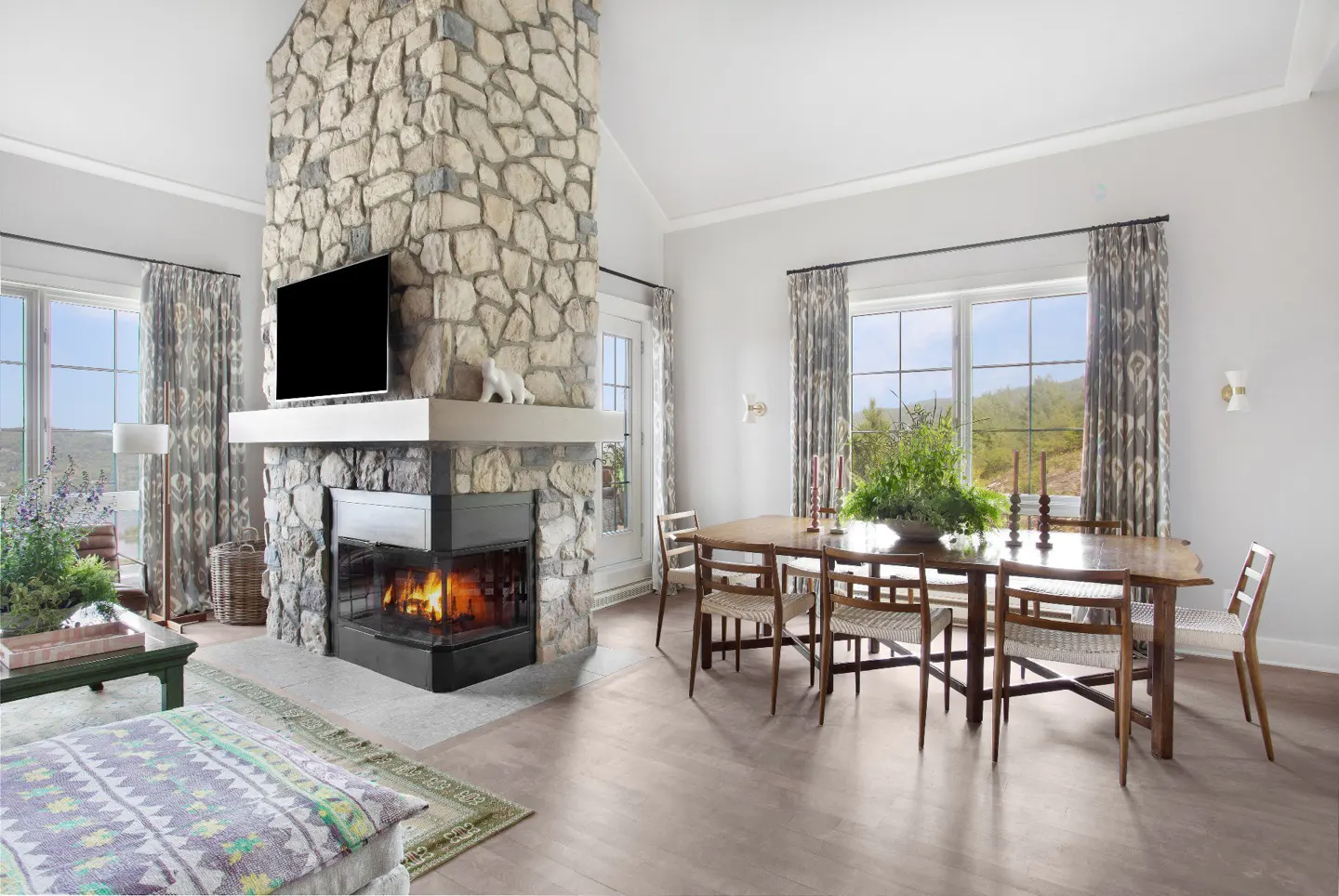 Living and dining area with stone fireplace, TV, and wood table with chairs. Large windows with patterned curtains.