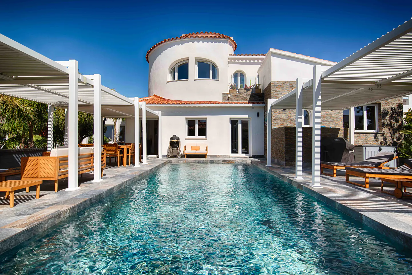 A long, narrow pool reflects a white house with a red tile roof under a bright blue sky. White pergolas line the pool's sides.