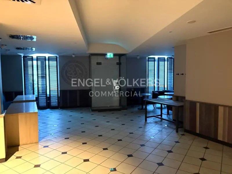 Empty commercial space with tile floors, tables, and shuttered windows. Engel & Völkers logo is visible on the wall.