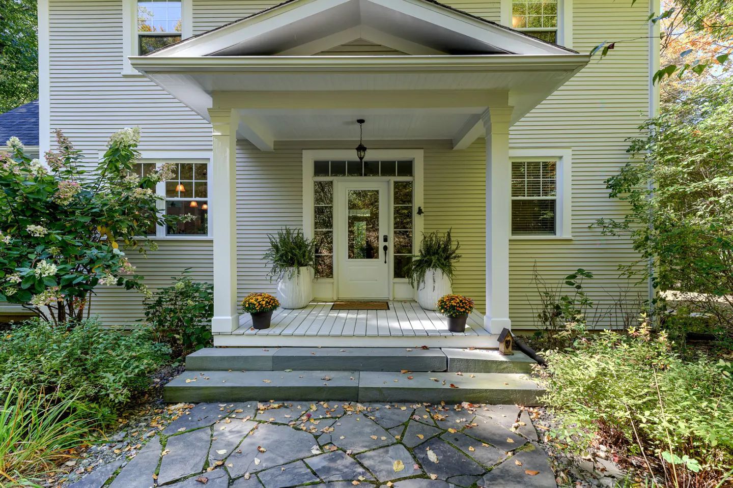 Front entrance of a light gray house with a white door and porch. Stone steps lead to the porch with potted plants and flowers.