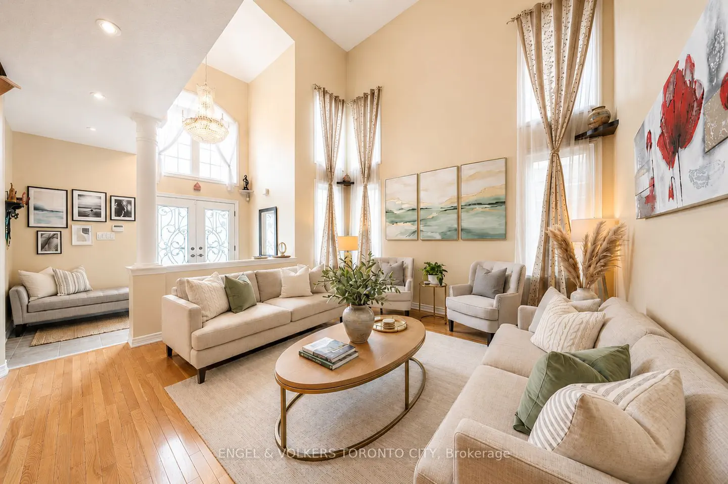 A bright, neutral living room with two sofas, armchairs, and a wooden coffee table on a light rug. Tall windows are draped with patterned curtains.