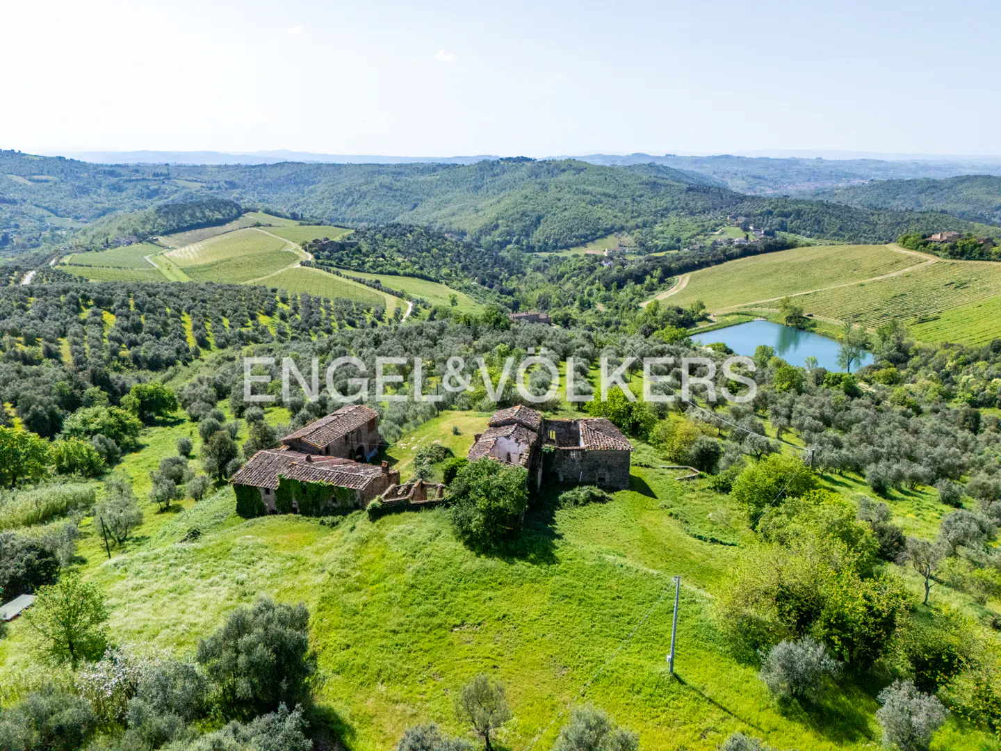 Aerial view of a Tuscan farmhouse with a pond, surrounded by olive groves and rolling green hills.