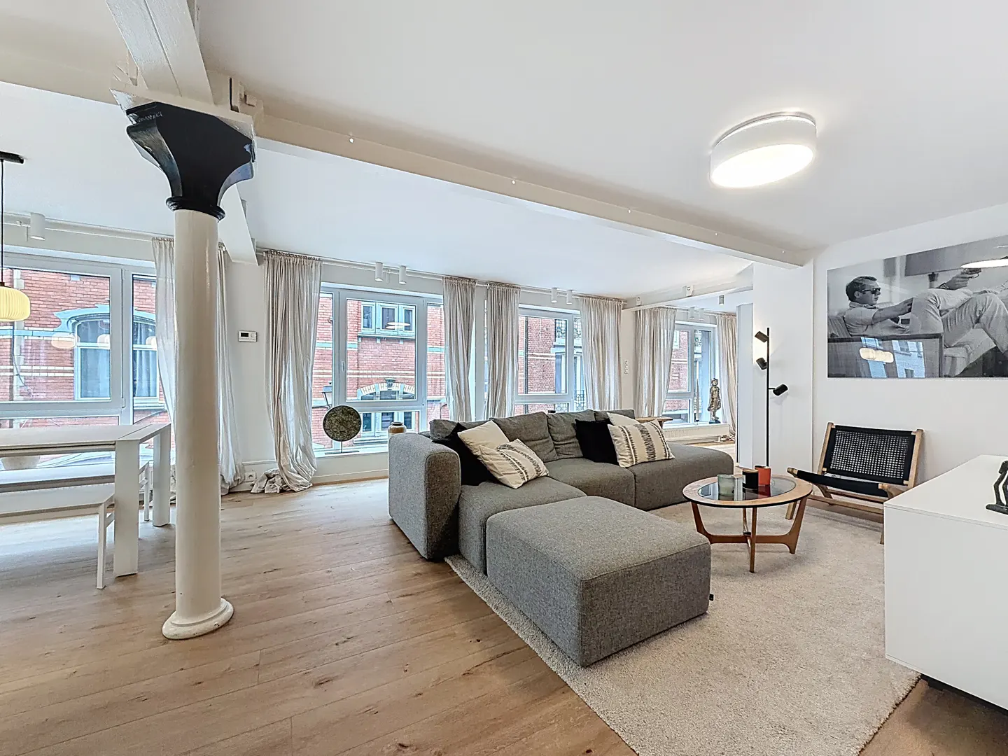 Bright, modern living room with wood floors, gray sectional sofa, and large windows with white curtains. A white column adds architectural detail.