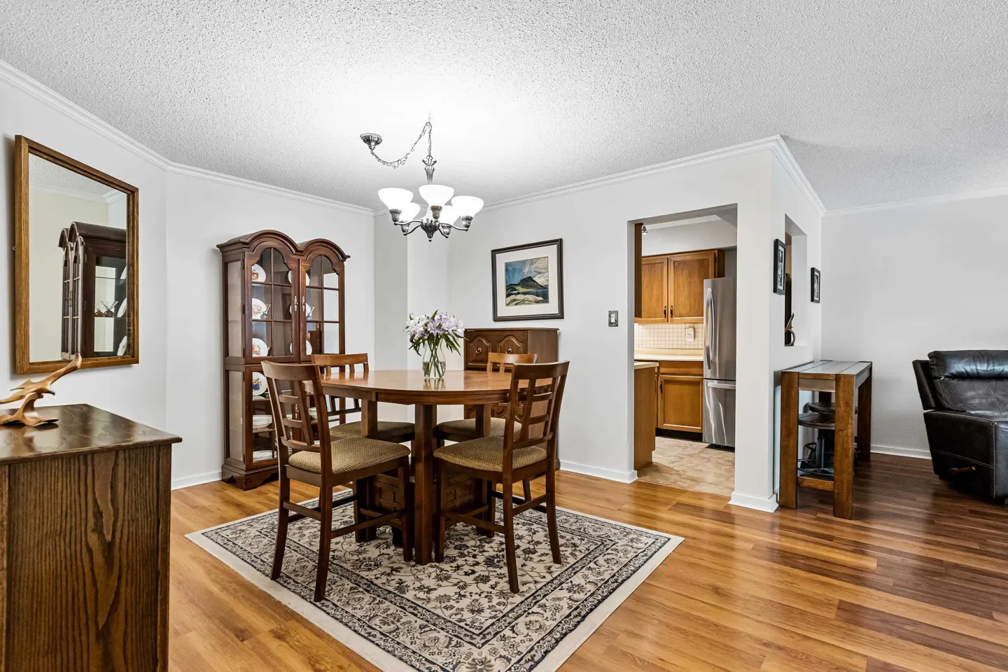 A dining room with wood floors, a round table with flowers, and a china cabinet. The kitchen is visible in the background.