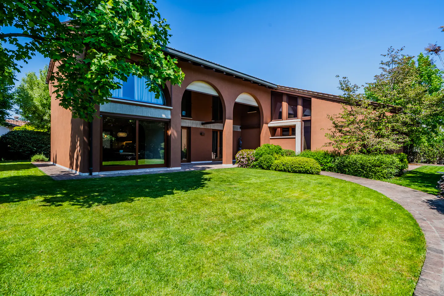 Exterior of a two-story brown house with arched entry, green lawn, trees, and blue sky.