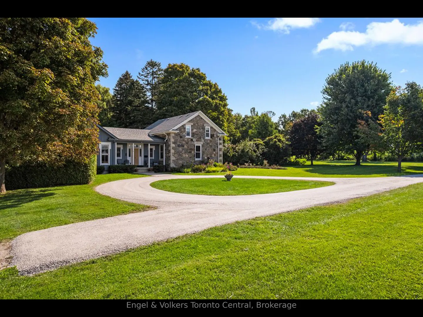 Exterior view of a stone house with a circular driveway, surrounded by green grass and trees under a blue sky.