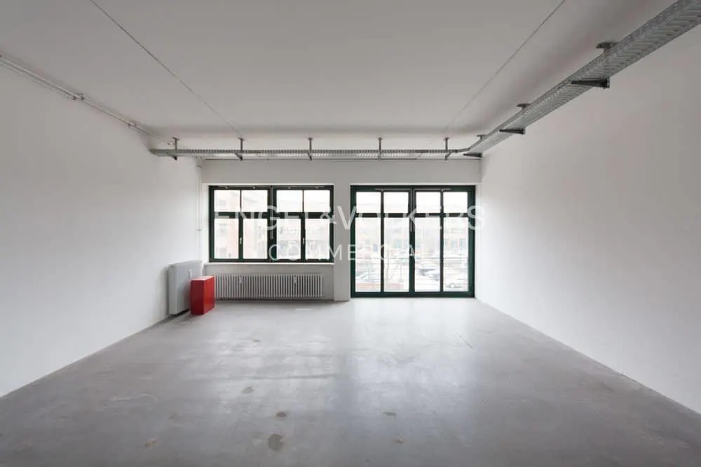 Empty room with white walls, gray concrete floor, and two large windows with dark green frames. A red radiator is visible.