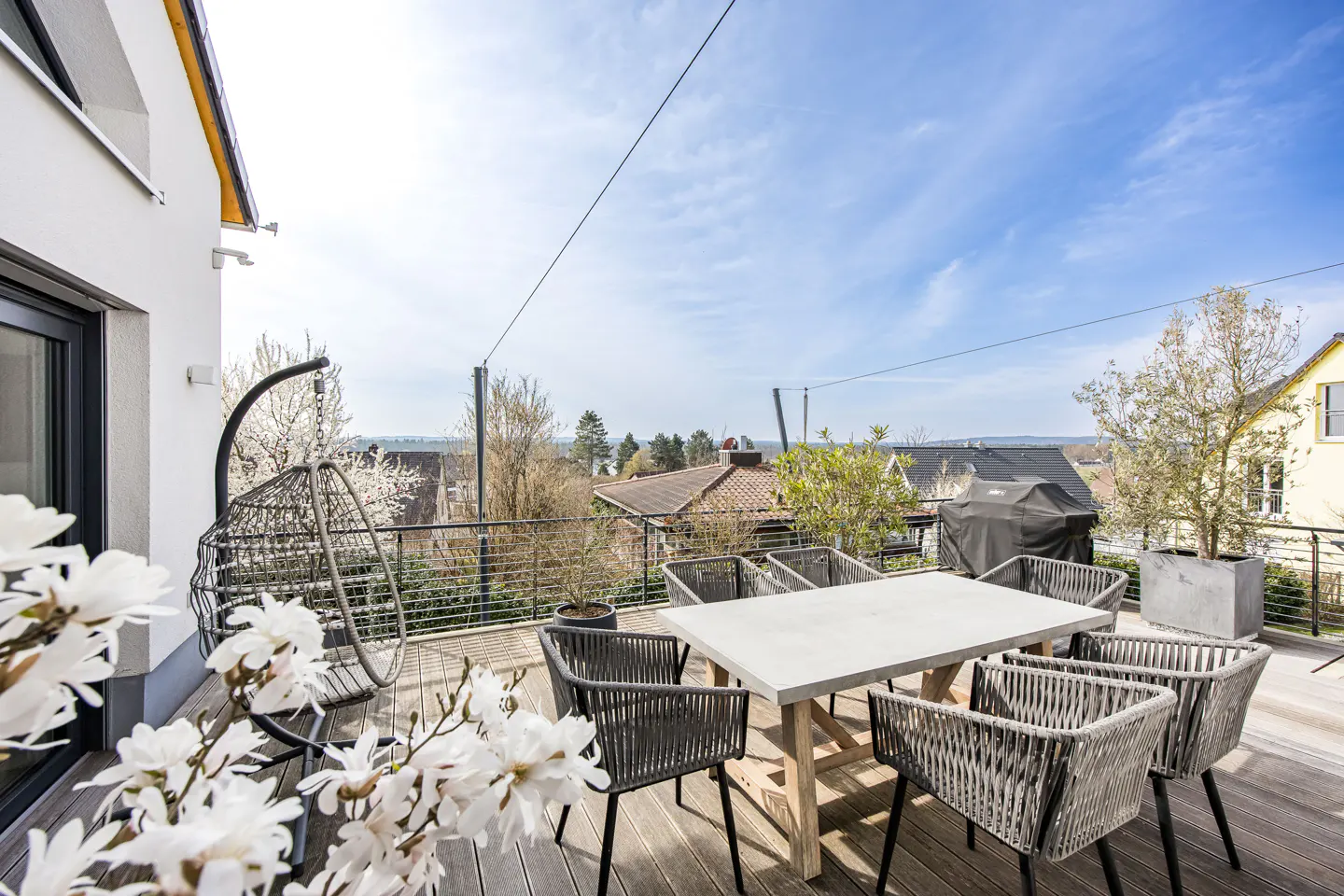 Outdoor patio with a table, chairs, and hanging swing chair on a wooden deck with a view of houses and trees under a blue sky.