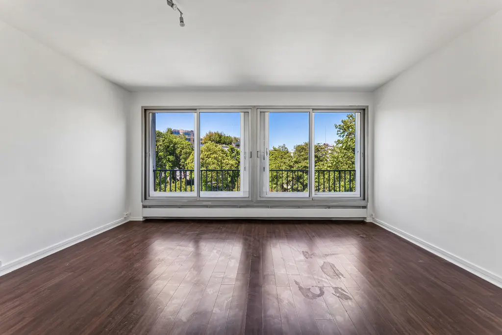 Empty room with dark wood floors, white walls, and large windows. Outside, trees and a blue sky are visible.