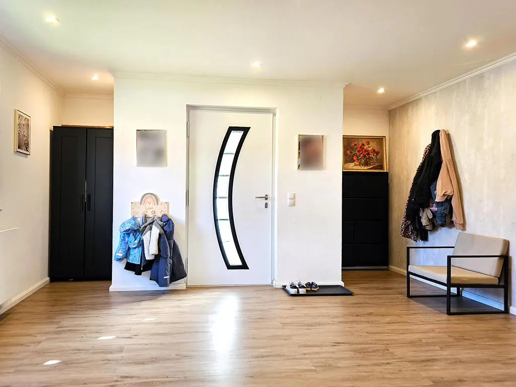 Entryway with wood floors, white walls, and recessed lighting. A white door with a black curved window is centered. A black wardrobe and a bench are visible.